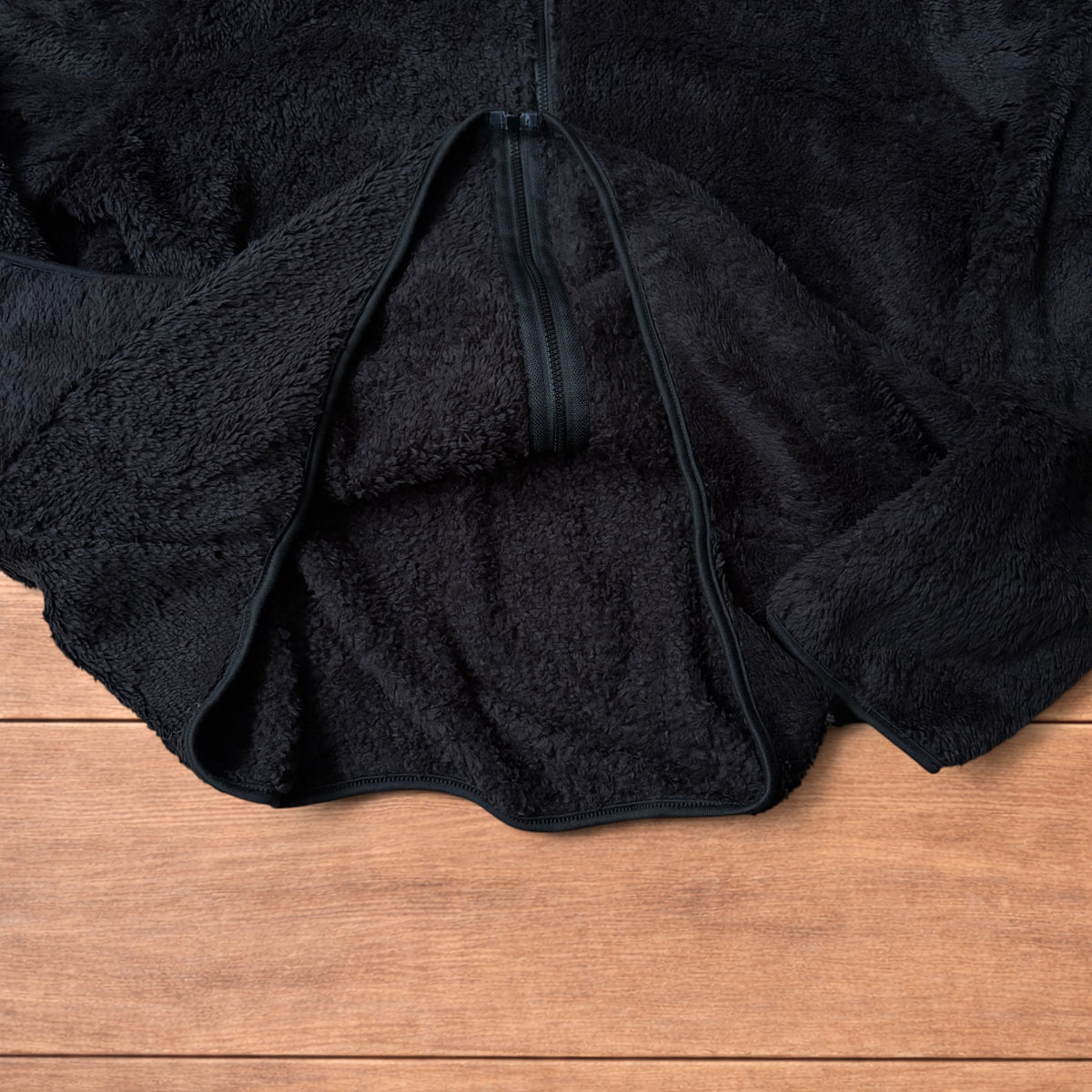 Black fleece fluffy zip up jacket laid flat on wooden floor showing full front, collar, sleeves, and pockets.