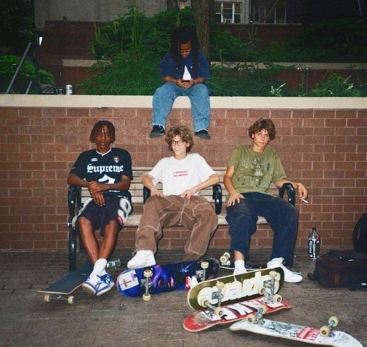 Group of skateborading teens sitting on a bench with their boards displayed in front of them.