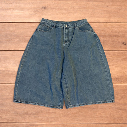 Mid blue baggy wide leg denim jorts shorts with front and back pockets laid flat on wooden background.
