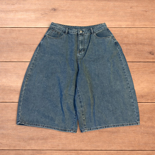 Mid blue baggy wide leg denim jorts shorts with front and back pockets laid flat on wooden background.