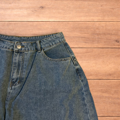 Mid blue baggy wide leg denim jorts shorts with front and back pockets laid flat on wooden background.