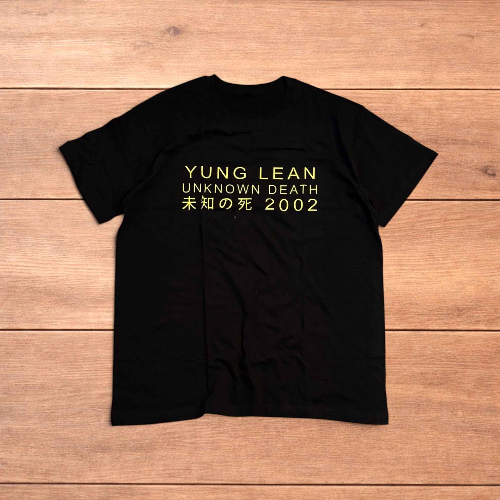 Black Yung Lean 2002 t-shirt with yellow front graphic text laid flat on a wooden surface.
