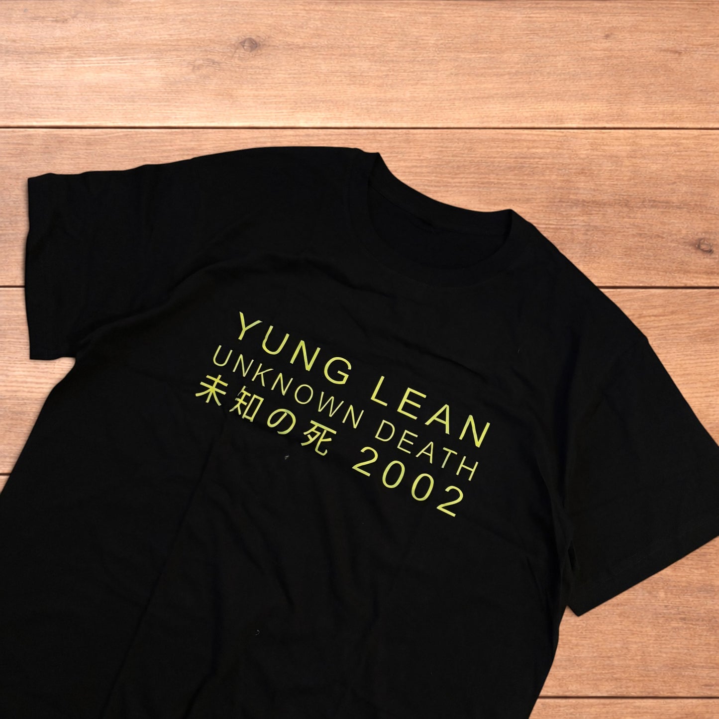 Black Yung Lean 2002 t-shirt with yellow front graphic text laid flat on a wooden surface.