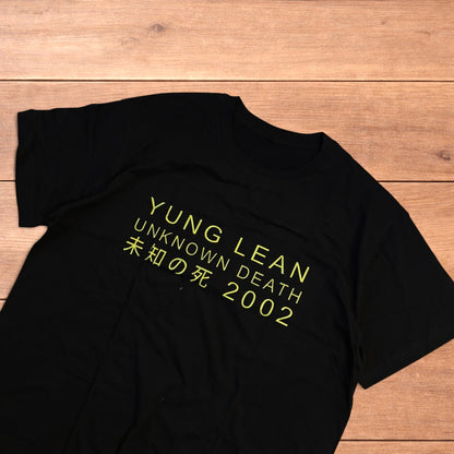 Black Yung Lean 2002 t-shirt with yellow front graphic text laid flat on a wooden surface.