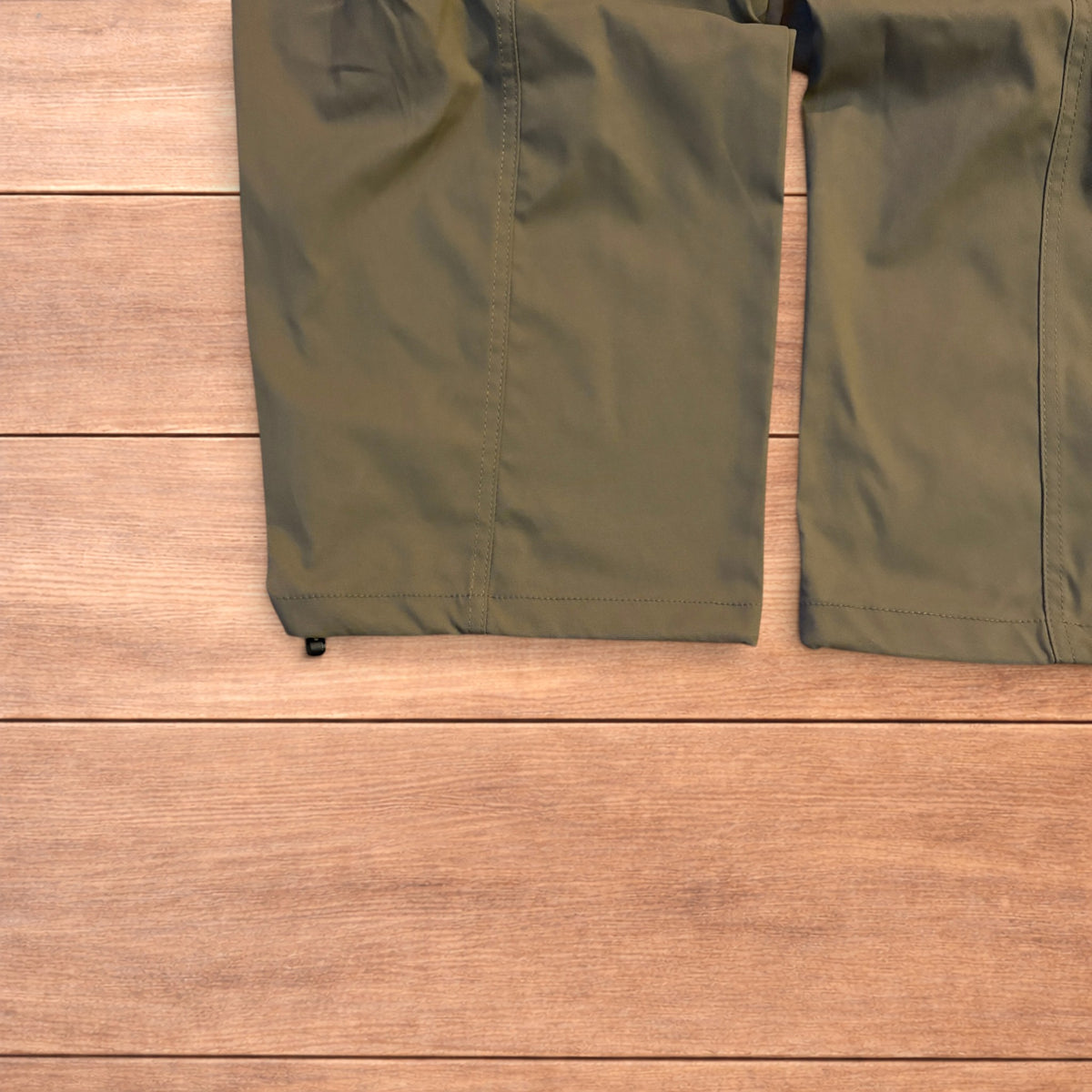 Khaki green baggy gorpcore tech hiking pants with drawstring waist and back flap pockets laid flat on wooden surface.