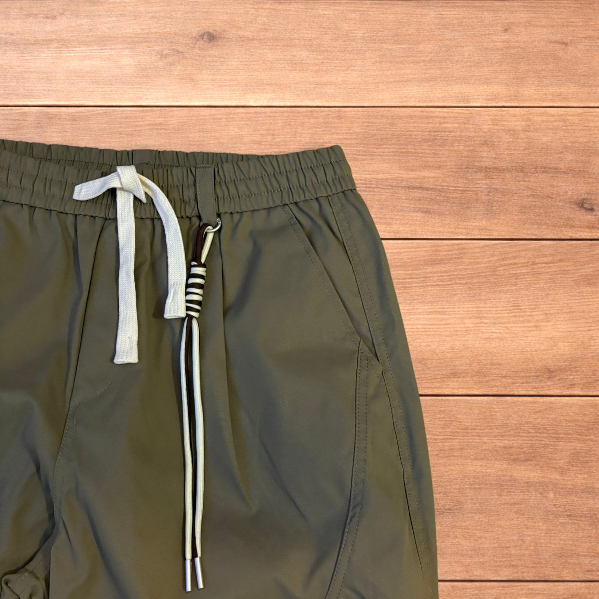 Khaki green baggy gorpcore tech hiking pants with drawstring waist and back flap pockets laid flat on wooden surface.