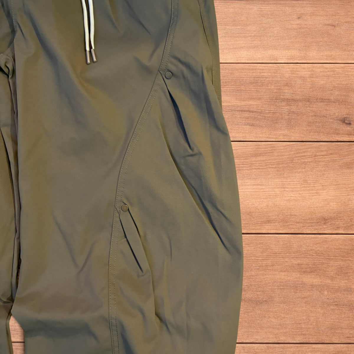 Khaki green baggy gorpcore tech hiking pants with drawstring waist and back flap pockets laid flat on wooden surface.