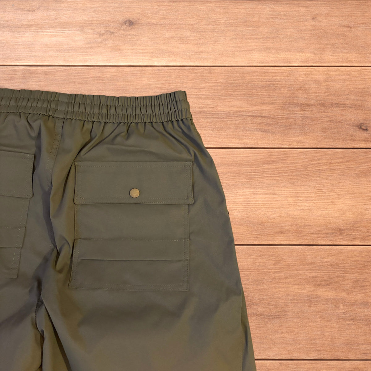 Khaki green baggy gorpcore tech hiking pants with drawstring waist and back flap pockets laid flat on wooden surface.