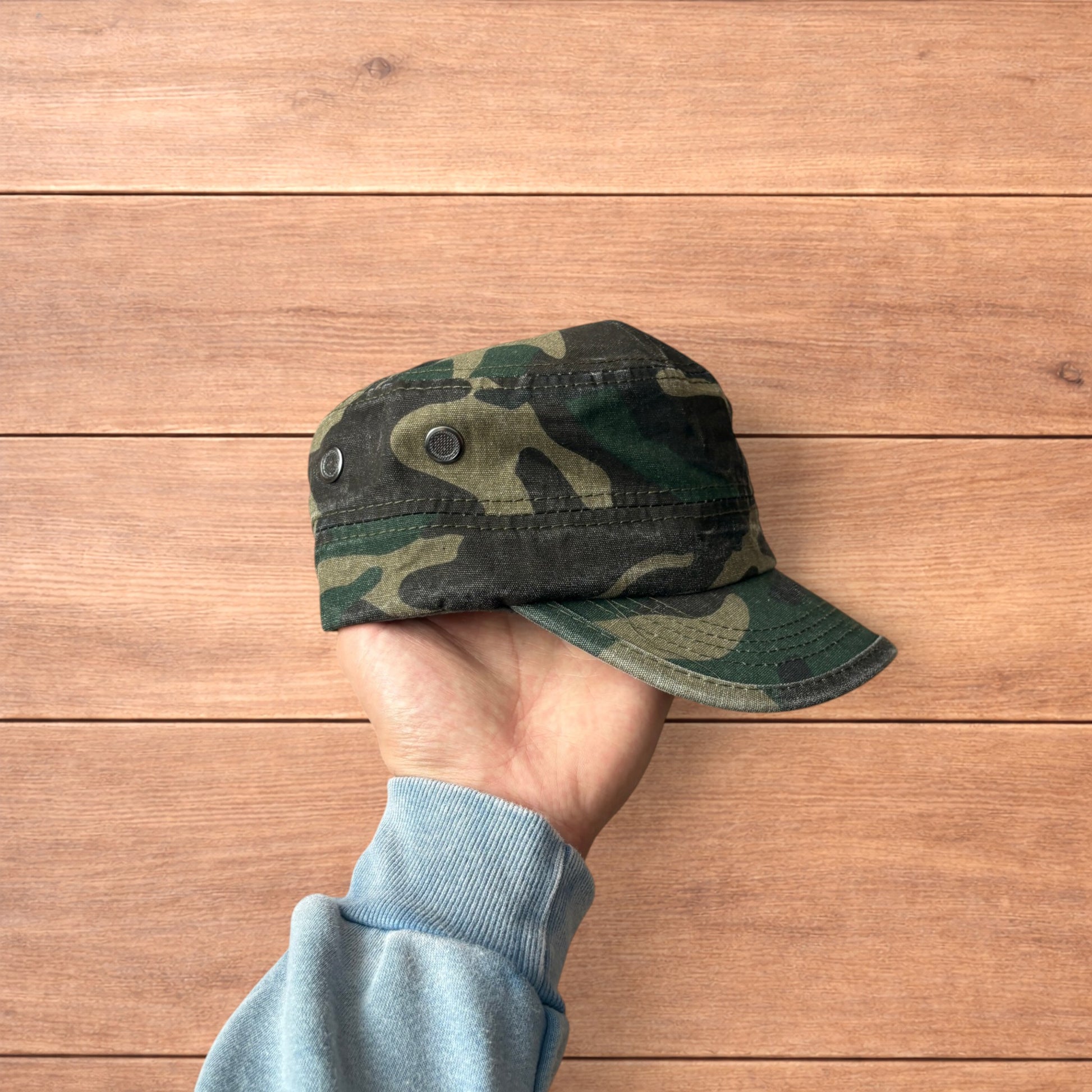Hand holding vintage style army camo cap hat with curved brim and side snap fasteners against wooden background.