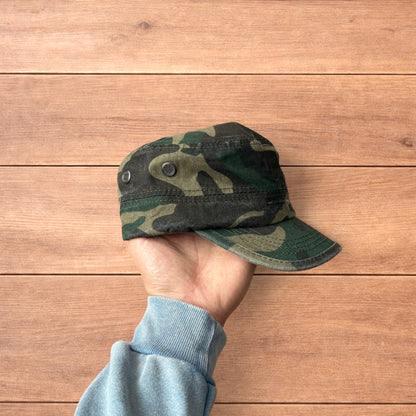 Hand holding vintage style army camo cap hat with curved brim and side snap fasteners against wooden background.