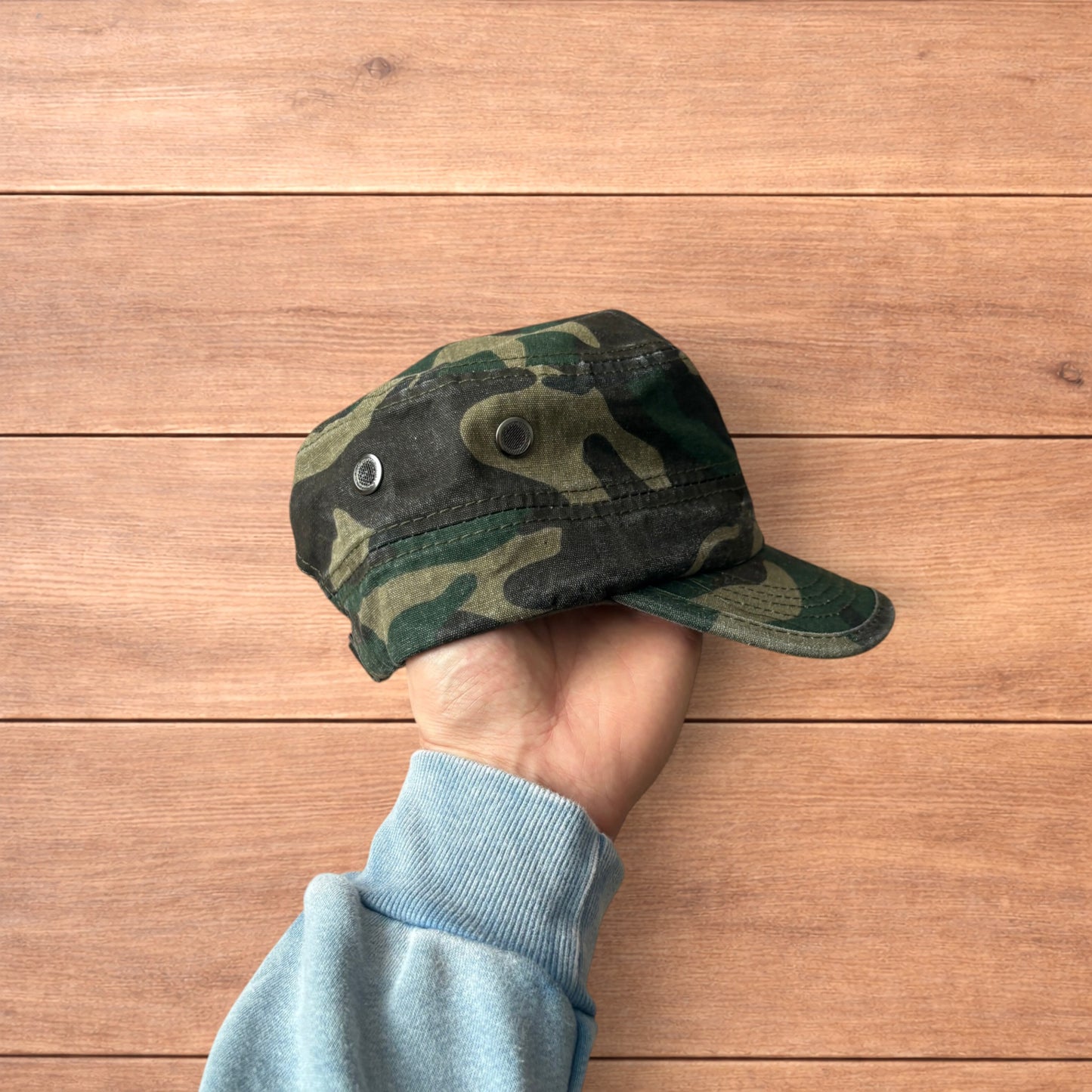 Hand holding vintage style army camo cap hat with curved brim and side snap fasteners against wooden background.