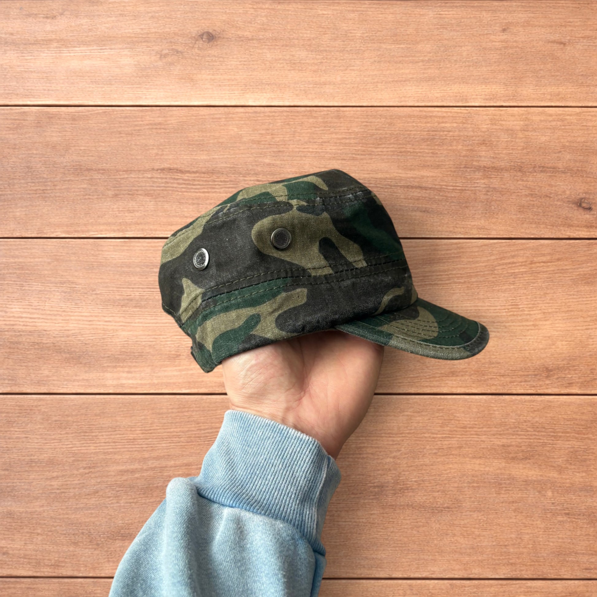 Hand holding vintage style army camo cap hat with curved brim and side snap fasteners against wooden background.