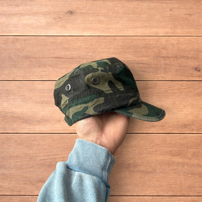 Hand holding vintage style army camo cap hat with curved brim and side snap fasteners against wooden background.