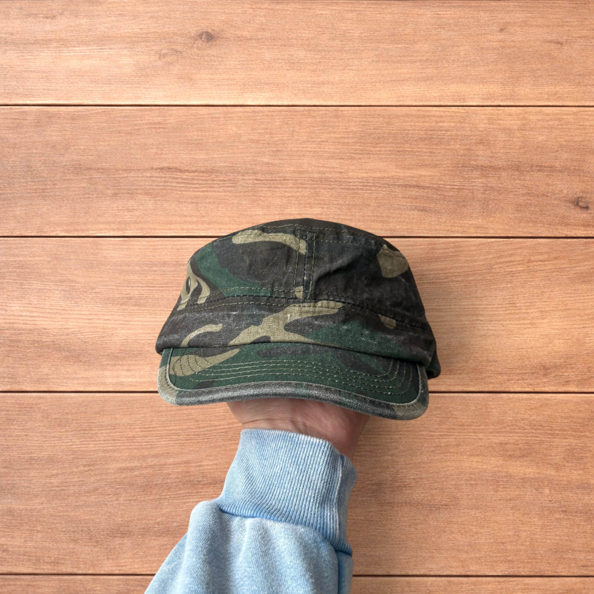 Hand holding vintage style army camo cap hat with curved brim and side snap fasteners against wooden background.