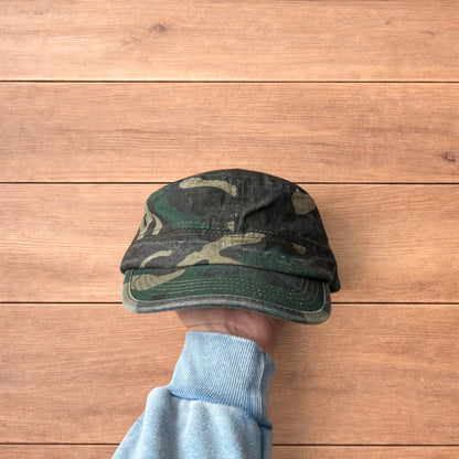 Hand holding vintage style army camo cap hat with curved brim and side snap fasteners against wooden background.