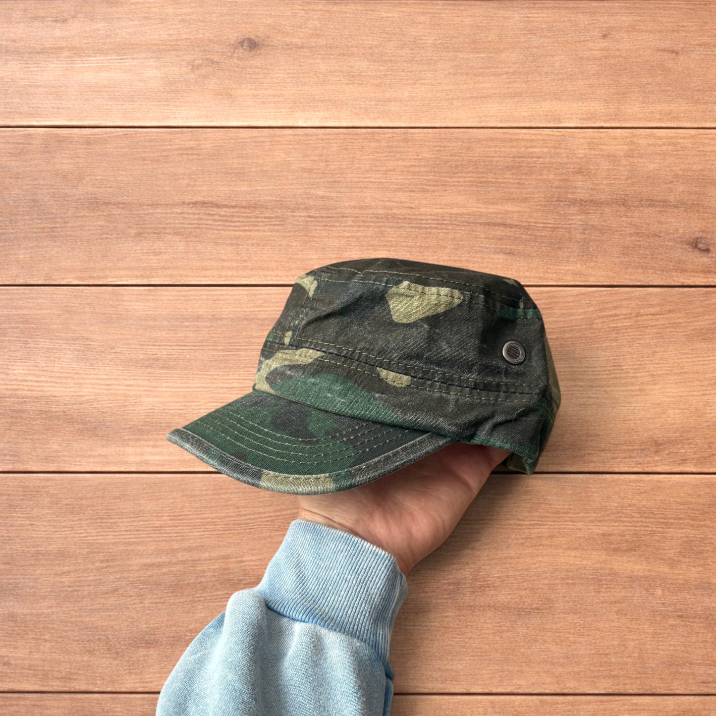Hand holding vintage style army camo cap hat with curved brim and side snap fasteners against wooden background.