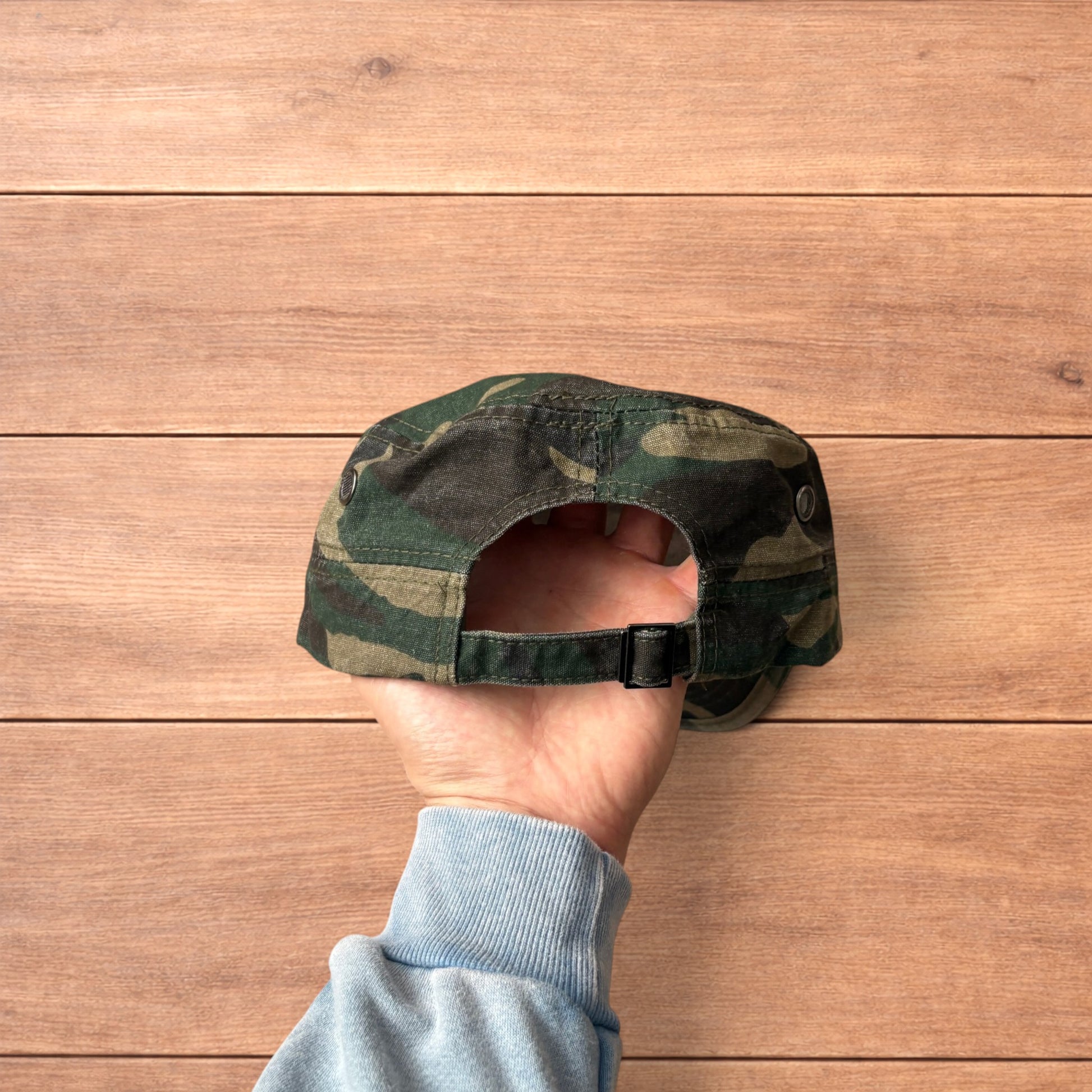 Hand holding vintage style army camo cap hat with curved brim and side snap fasteners against wooden background.