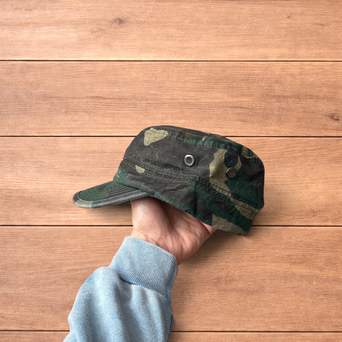 Hand holding vintage style army camo cap hat with curved brim and side snap fasteners against wooden background.