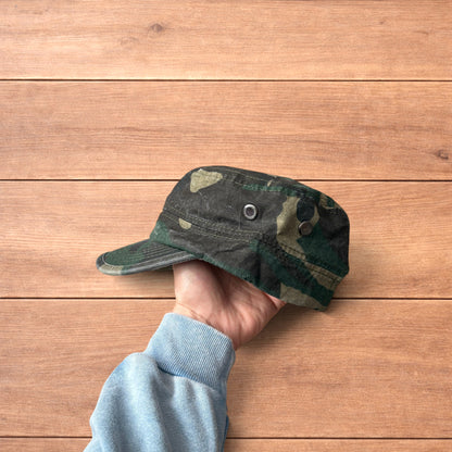 Hand holding vintage style army camo cap hat with curved brim and side snap fasteners against wooden background.