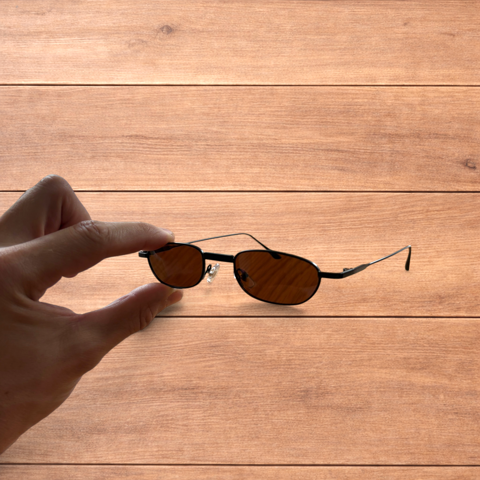 Hand holding slim vintage style sunglasses with black frame and brown lenses over wooden background.