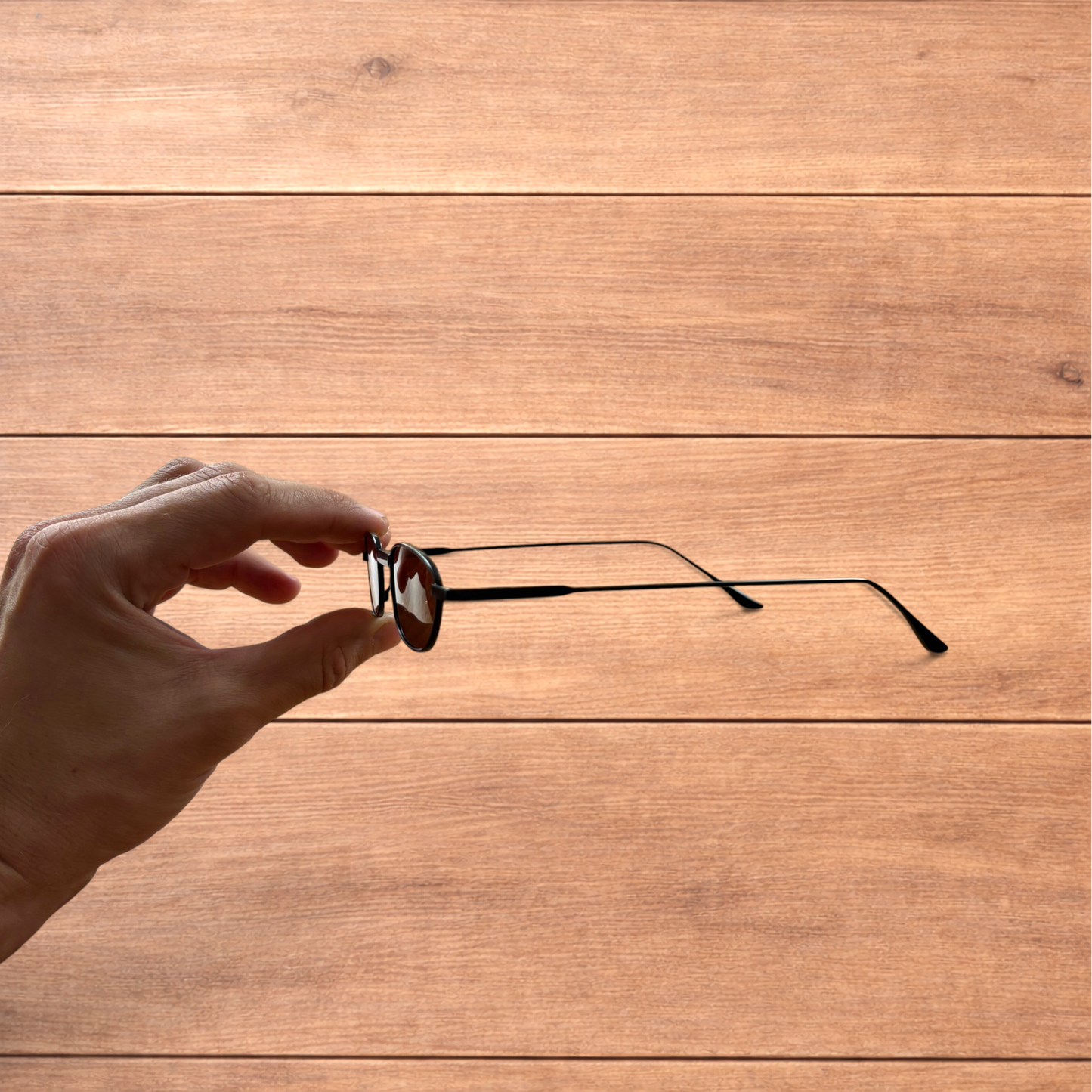 Hand holding slim vintage style sunglasses with black frame and brown lenses over wooden background.