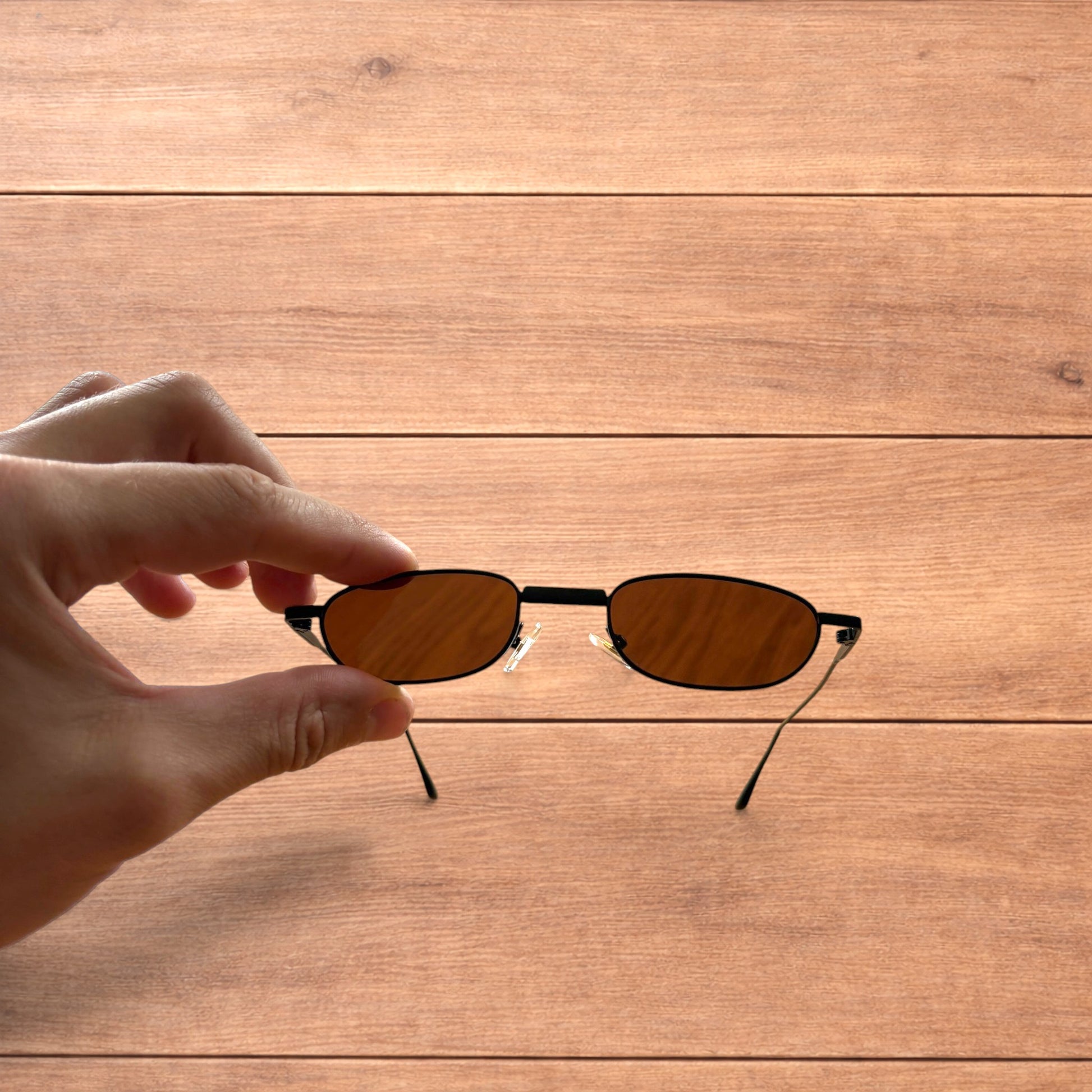 Hand holding slim vintage style sunglasses with black frame and brown lenses over wooden background.