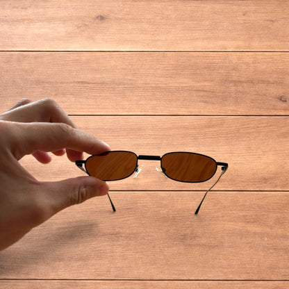 Hand holding slim vintage style sunglasses with black frame and brown lenses over wooden background.
