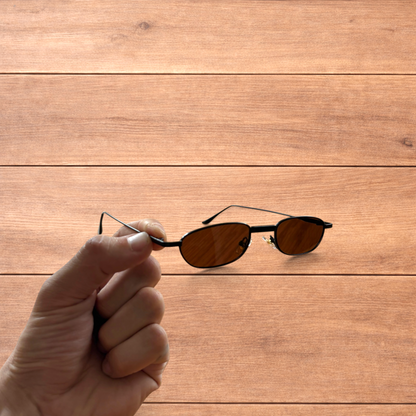 Hand holding slim vintage style sunglasses with black frame and brown lenses over wooden background.