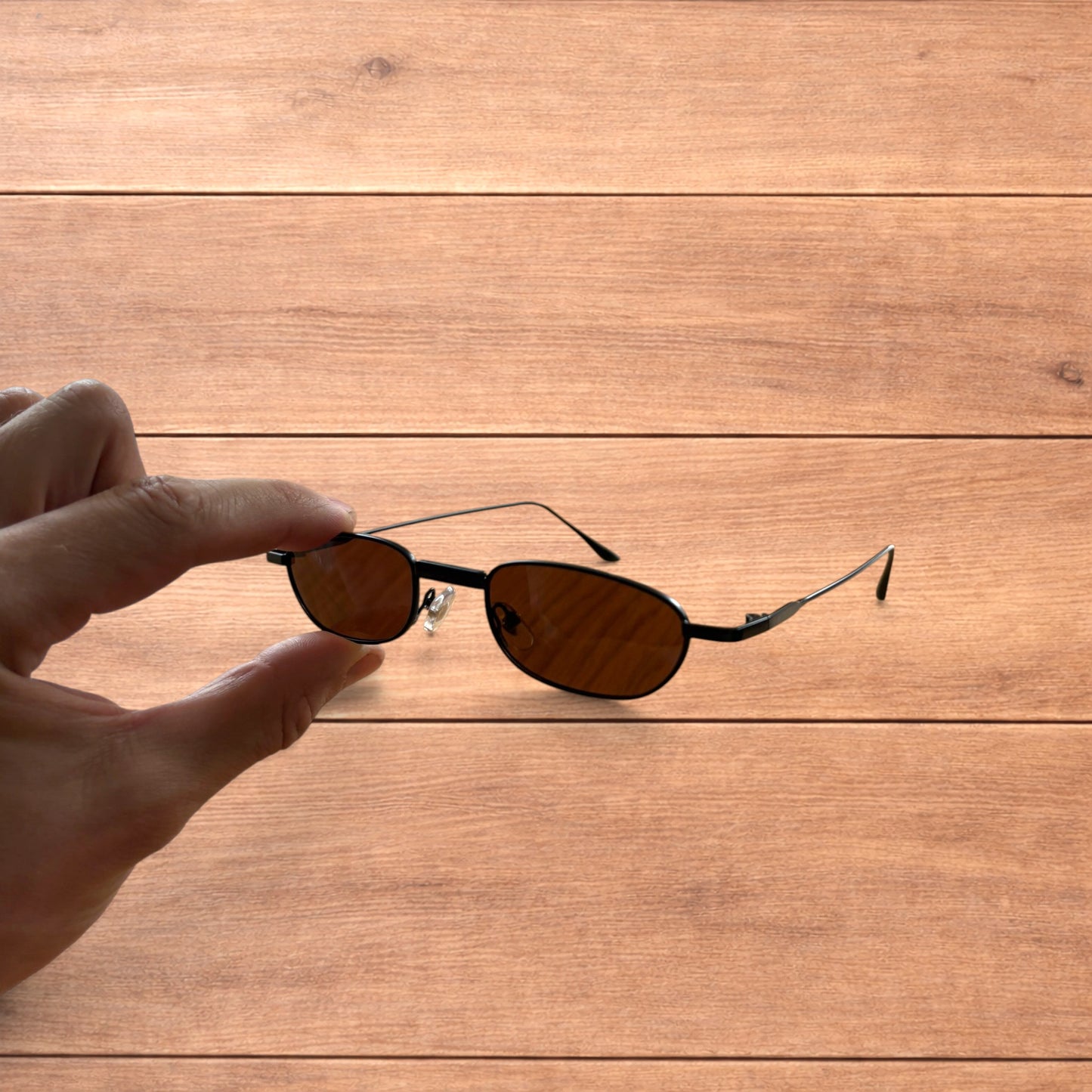 Hand holding slim vintage style sunglasses with black frame and brown lenses over wooden background.