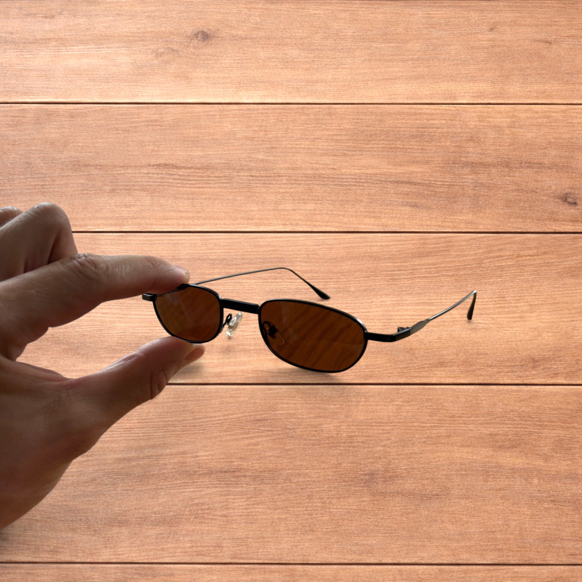 Hand holding slim vintage style sunglasses with black frame and brown lenses over wooden background.