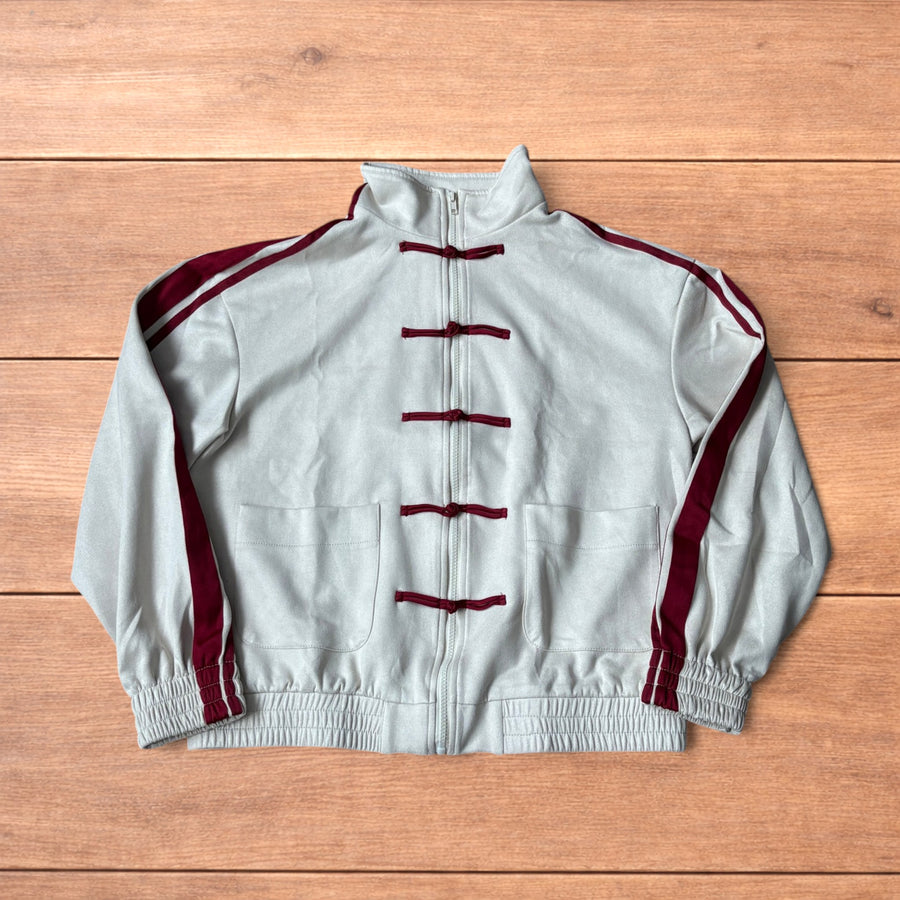 Cream Japanese style zip up track jacket with maroon sleeve stripes and knot button detailing laid flat on wooden surface.