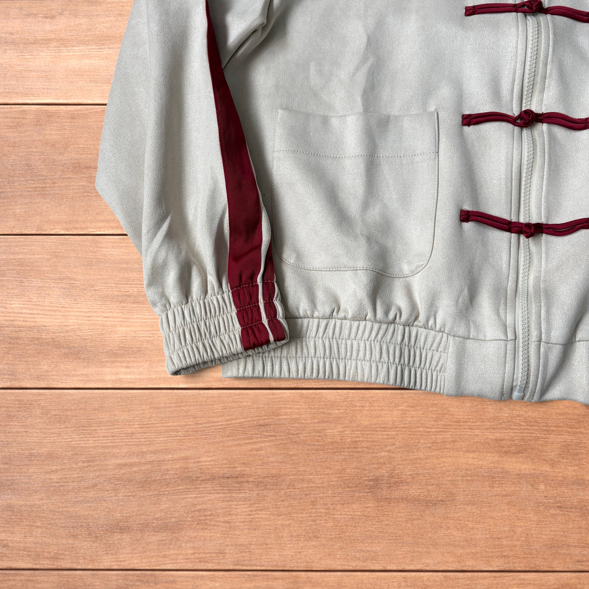 Cream Japanese style zip up track jacket with maroon sleeve stripes and knot button detailing laid flat on wooden surface.
