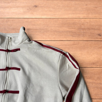 Cream Japanese style zip up track jacket with maroon sleeve stripes and knot button detailing laid flat on wooden surface.