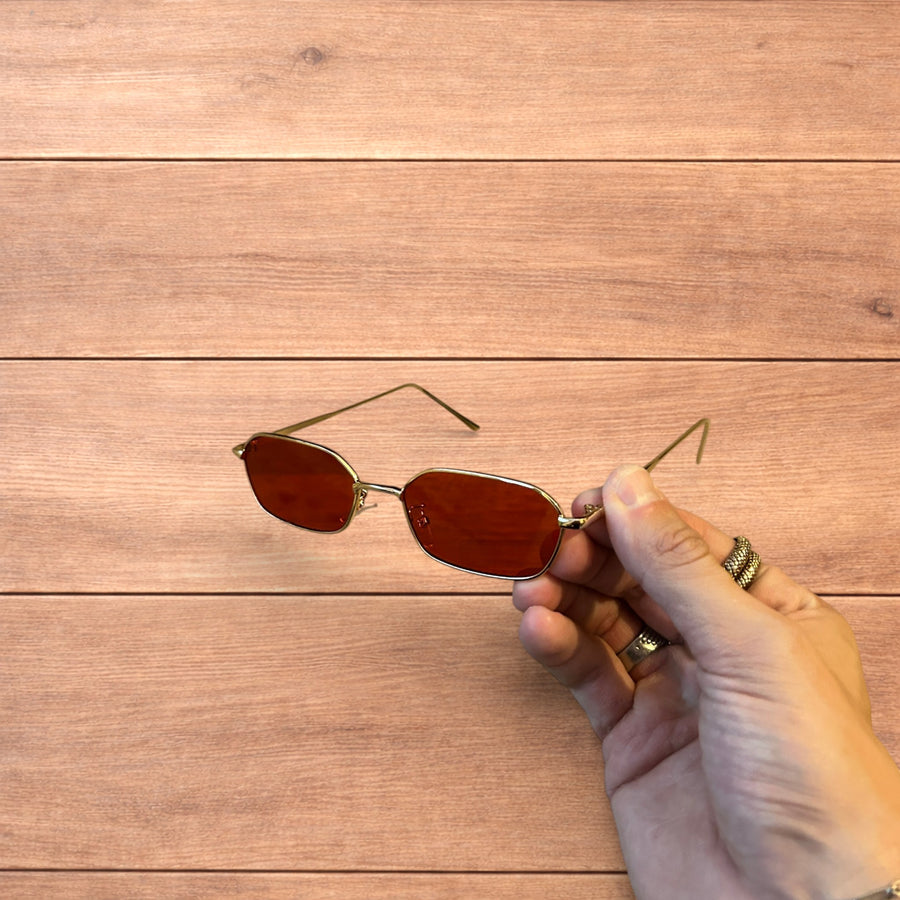 Hand holding gold metal frame sunglasses with red polarised rectangular lenses over a wooden background.