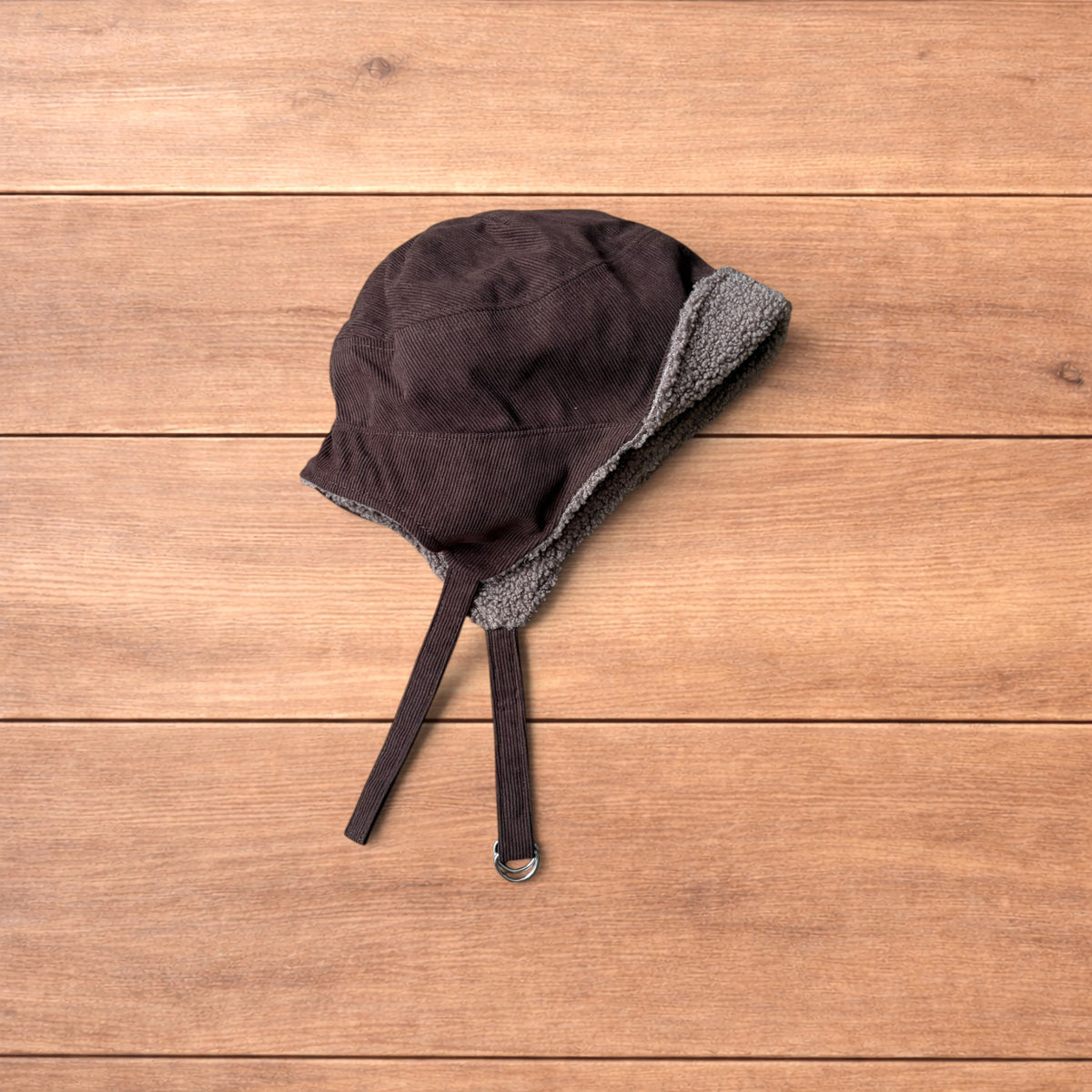 Brown Korean trapper hat with corduroy outer and fluffy woolen lining, laid flat on wooden surface showing ear flaps and chin strap.