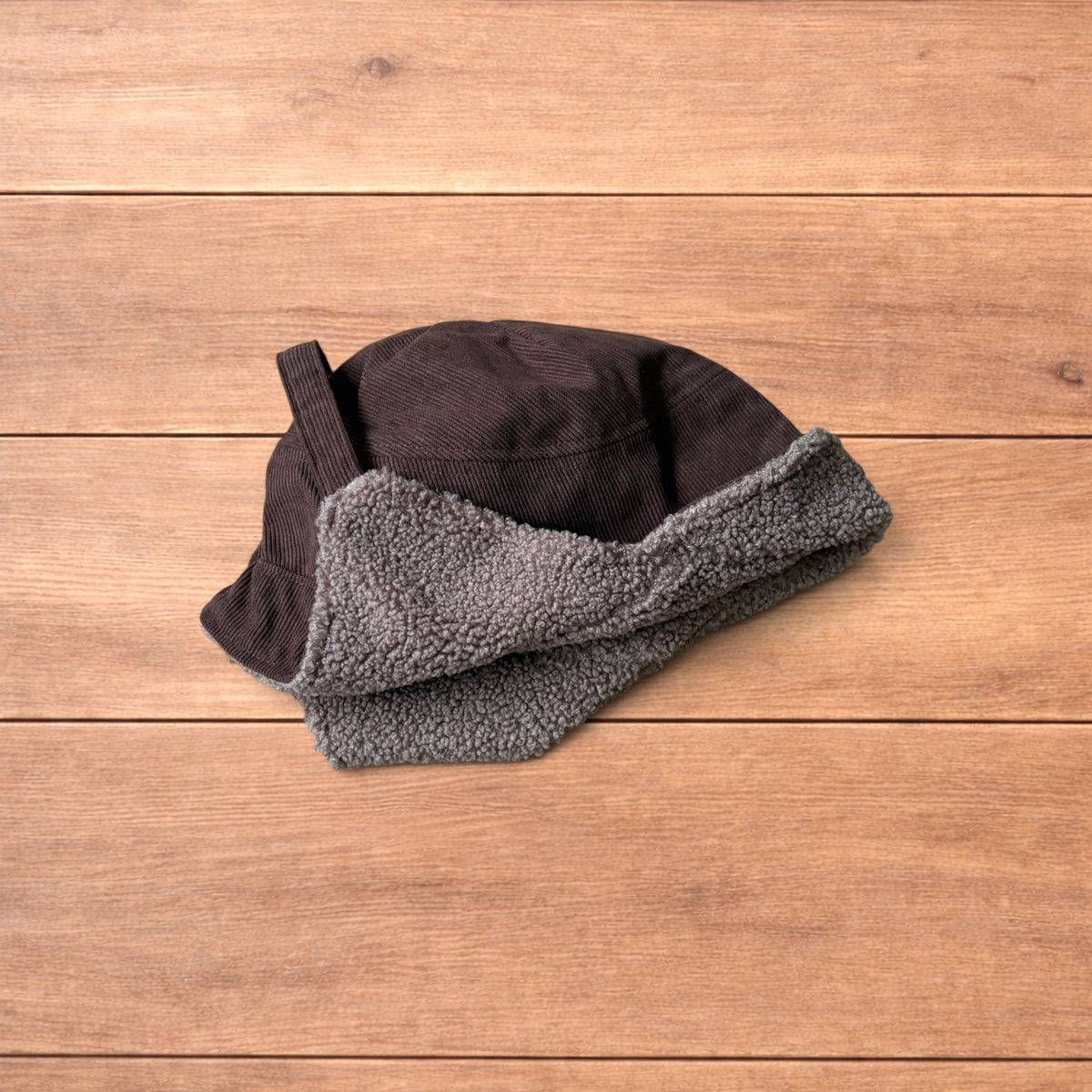 Brown Korean trapper hat with corduroy outer and fluffy woolen lining, laid flat on wooden surface showing ear flaps and chin strap.