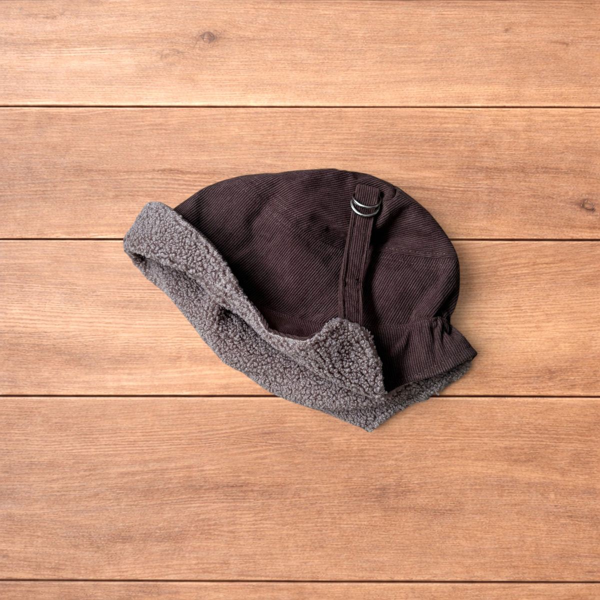 Brown Korean trapper hat with corduroy outer and fluffy woolen lining, laid flat on wooden surface showing ear flaps and chin strap.