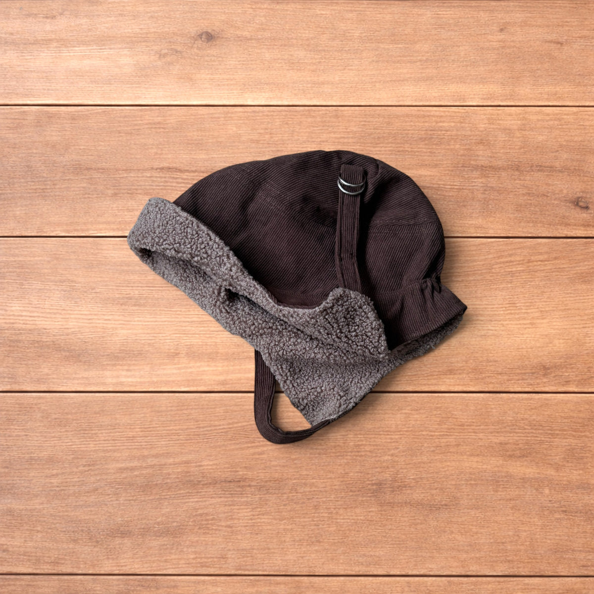 Brown Korean trapper hat with corduroy outer and fluffy woolen lining, laid flat on wooden surface showing ear flaps and chin strap.