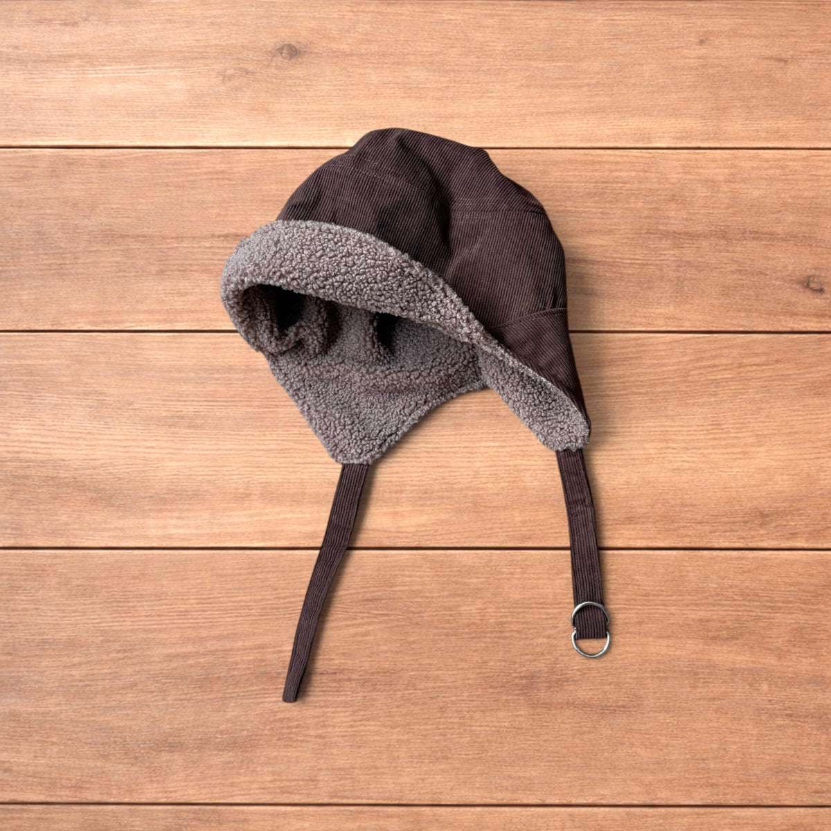 Brown Korean trapper hat with corduroy outer and fluffy woolen lining, laid flat on wooden surface showing ear flaps and chin strap.