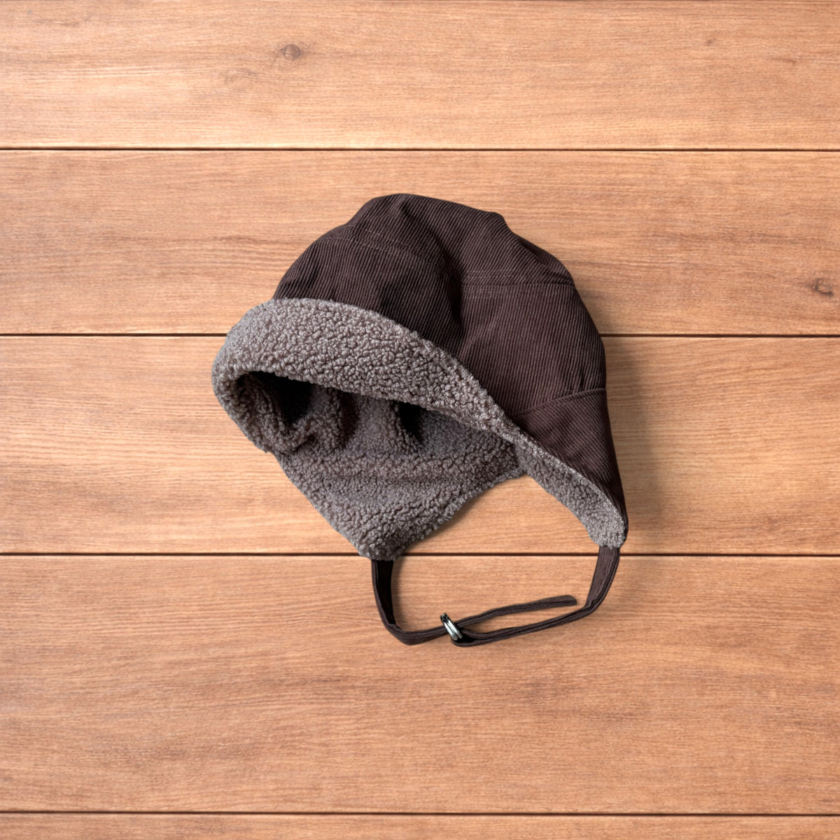 Brown Korean trapper hat with corduroy outer and fluffy woolen lining, laid flat on wooden surface showing ear flaps and chin strap.