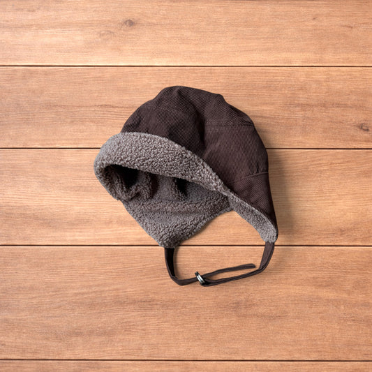Brown Korean trapper hat with corduroy outer and fluffy woolen lining, laid flat on wooden surface showing ear flaps and chin strap.