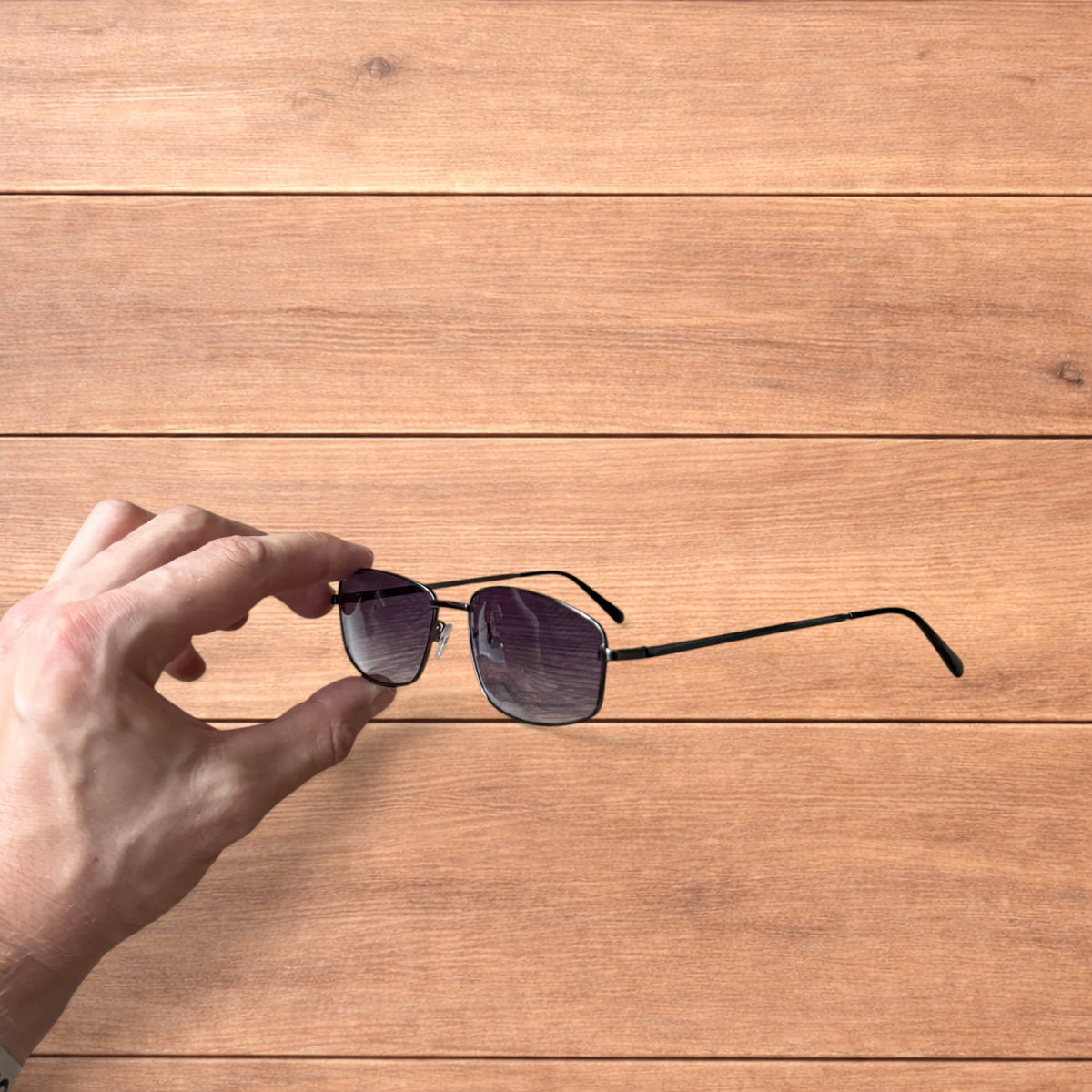 Hand holding vintage style black metal frame sunglasses with purple lenses against a wooden background.