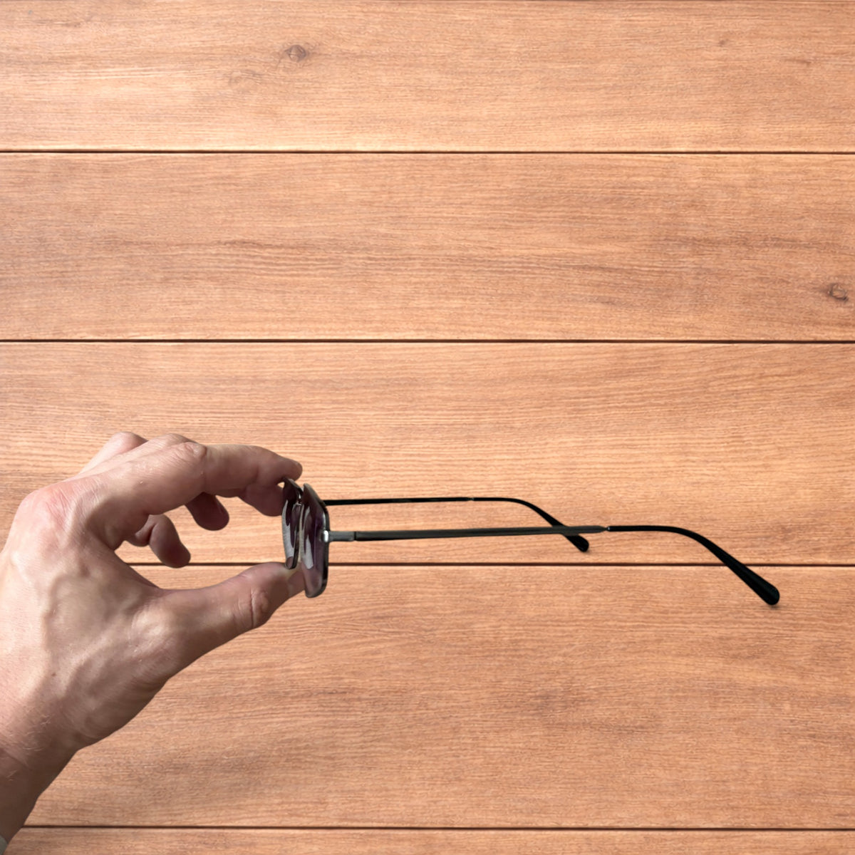 Hand holding vintage style black metal frame sunglasses with purple lenses against a wooden background.