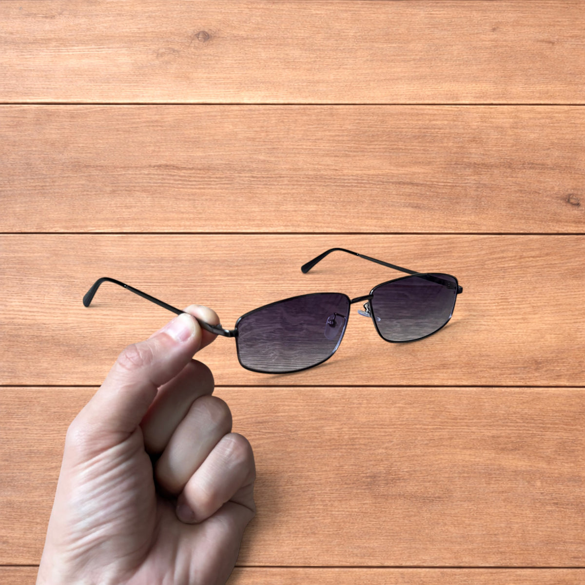 Hand holding vintage style black metal frame sunglasses with purple lenses against a wooden background.