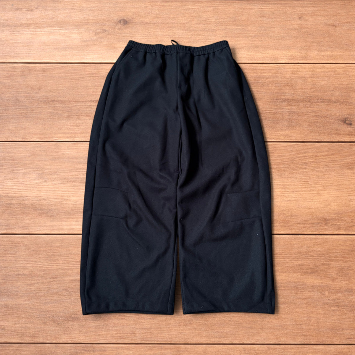 Black baggy wool trousers laid flat on wooden floor showing elasticated waistband and pockets.