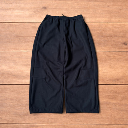 Black baggy wool trousers laid flat on wooden floor showing elasticated waistband and pockets.