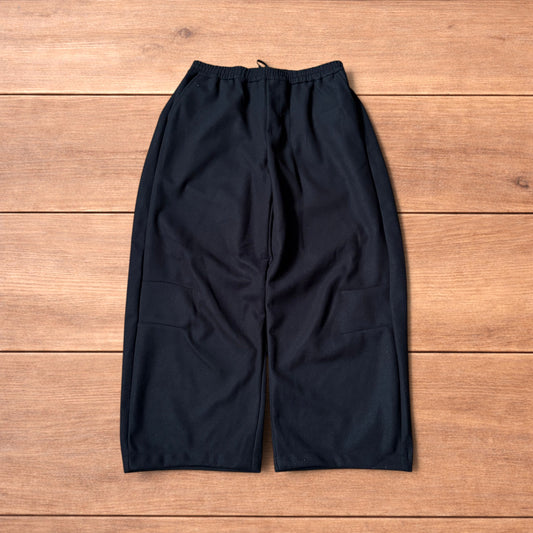 Black baggy wool trousers laid flat on wooden floor showing elasticated waistband and pockets.