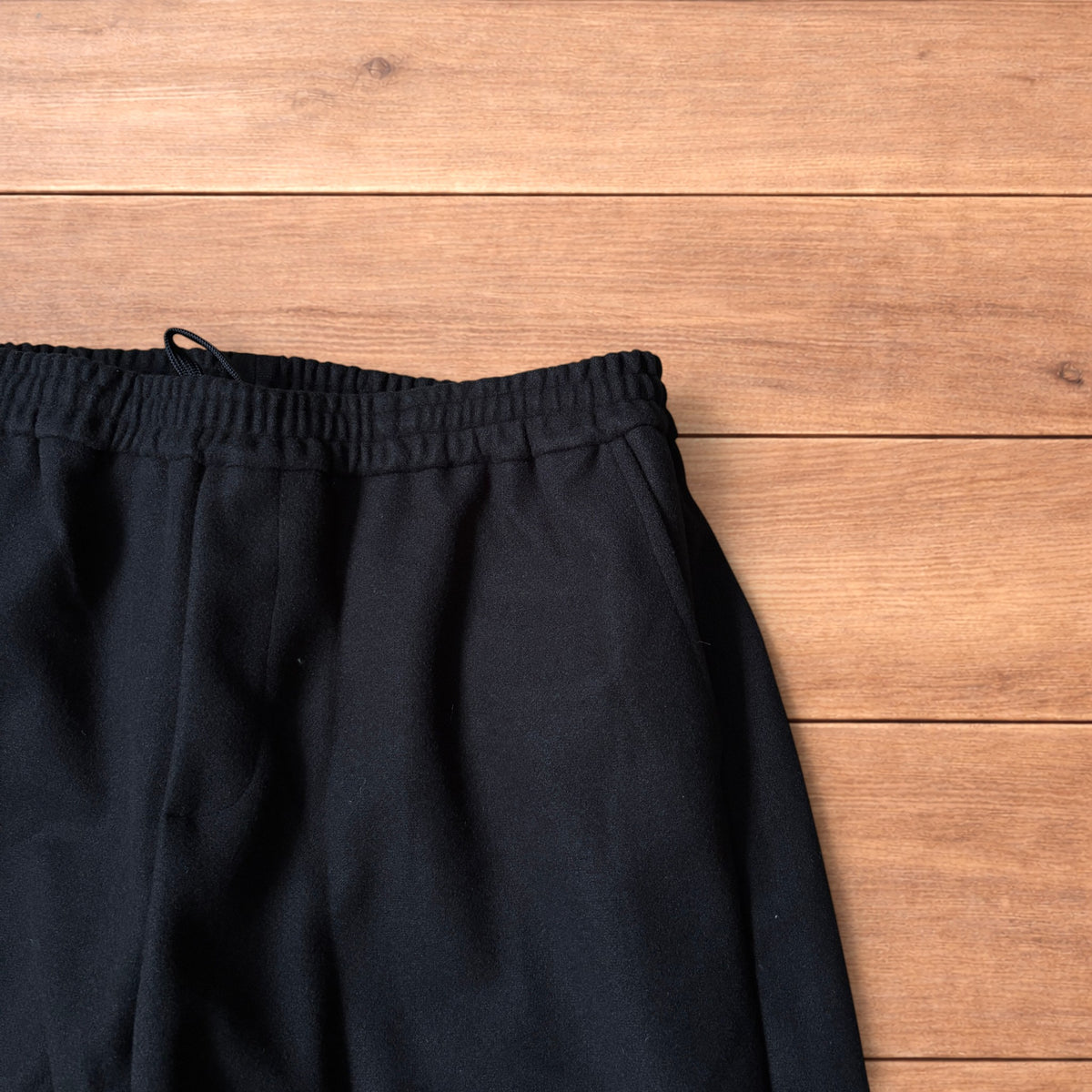 Black baggy wool trousers laid flat on wooden floor showing elasticated waistband and pockets.