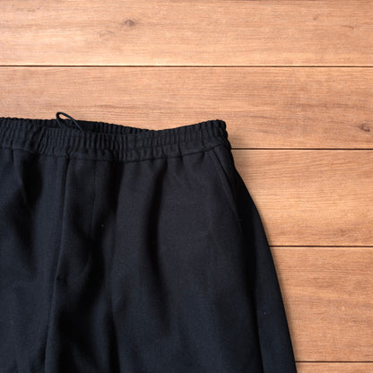 Black baggy wool trousers laid flat on wooden floor showing elasticated waistband and pockets.