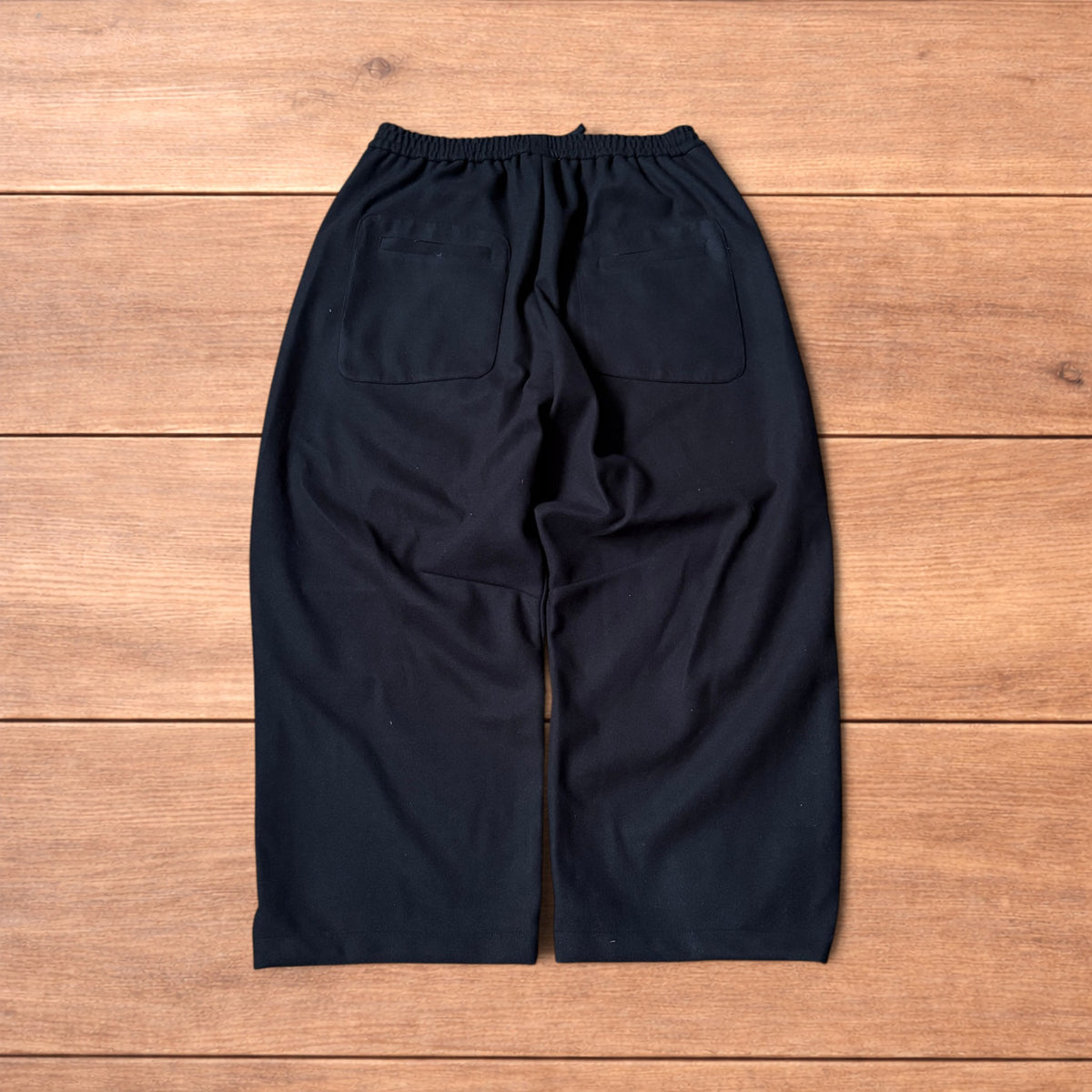 Black baggy wool trousers laid flat on wooden floor showing elasticated waistband and pockets.